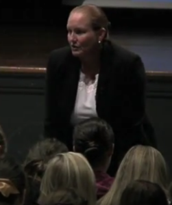A woman speaking to an audience in a dark setting.